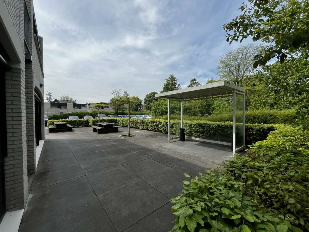 Moderne, betegelde buitenruimte aan de Burgemeester Verderlaan met zitbankjes, overkapping en groene beplanting.